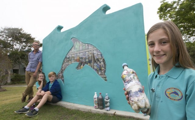 Part of the Ocean Shores School’s Eco Warrior team, Mackenzie Ormiston, Louis Lamont and Mia Shaw –along with teacher Wayne Gibbons – show the rendered wall they built from plastic milk bottles, filled with used plastic. Photo Jeff ‘All Plastic Since 1986’ Dawson