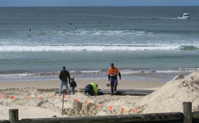 Contractors work on the spoil dump on the beach, while surfers enjoy the surf just offshore.